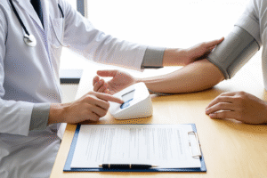 A healthcare professional uses a digital blood pressure monitor to check a patient’s blood pressure in a well-lit medical office, with a clipboard and pen on the table.